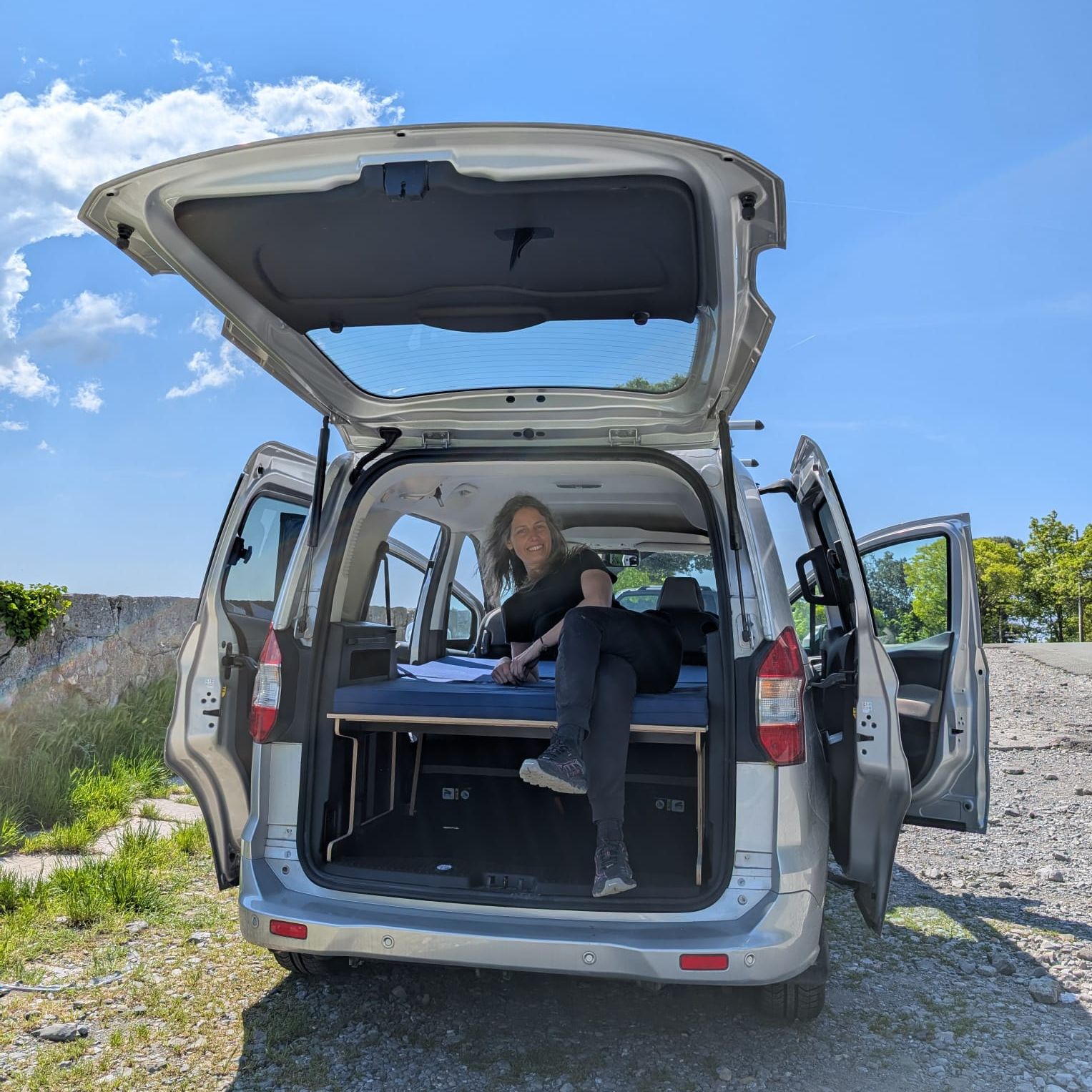 Smiling person sits in the rear of an open-back van on a bed platform, with doors wide open under a clear blue sky.